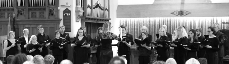 Group of singers dressed in black, holding black folders, standing in a church building, conducted by a female conductor.